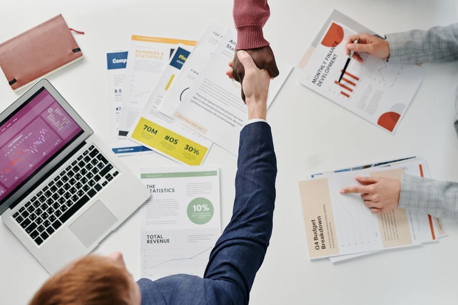 Business professionals engaging in a handshake over a desk filled with financial documents and a laptop.